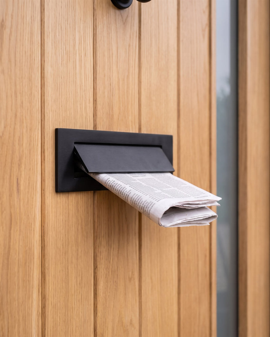 Matt black stainless steel letter plate on an oak boarded front door with a newspaper in the flap