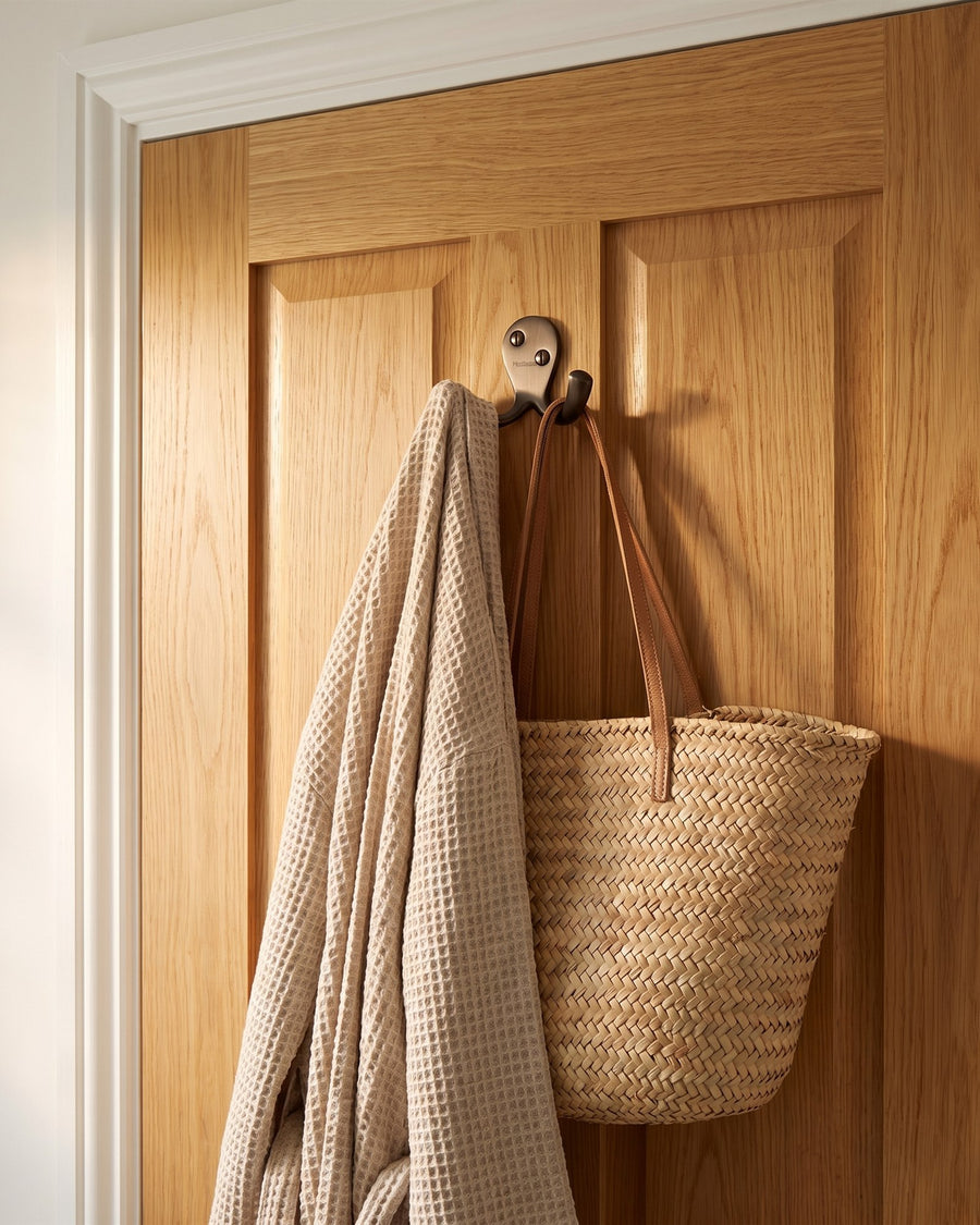 Matt bronze double robe hook holding a linen robe and a woven bag on a natural oak door