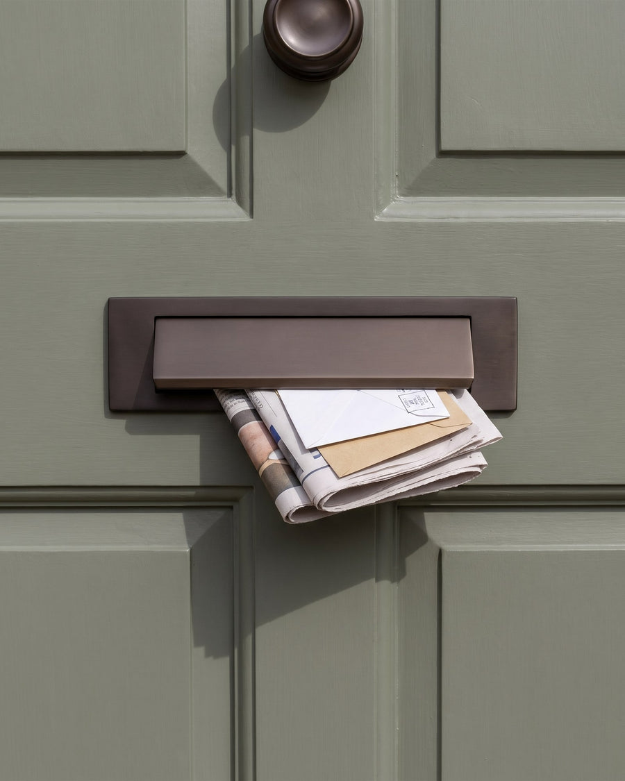 Matt bronze gravity flap letter plate with a newspaper and letters being delivered on a sage green front door