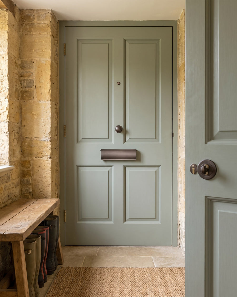 Inside of the sage green front door with a matt bronze letter tidy, in a stone-walled hall with limestone floor, seagrass mat and boots under an oak bench