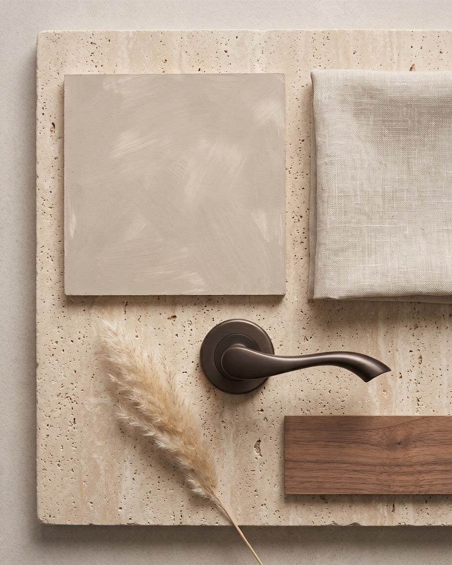 Flat-lay of limewash plaster, oatmeal linen, walnut veneer, dried pampas grass and a matt bronze Algarve lever on rose on honed travertine
