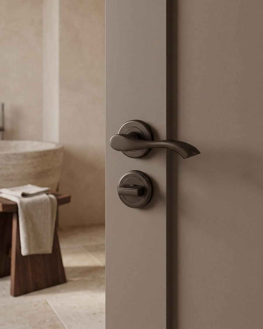 Mushroom-painted bathroom door with a matt bronze Algarve lever on rose and matching turn and release, travertine floor and stone basin beyond