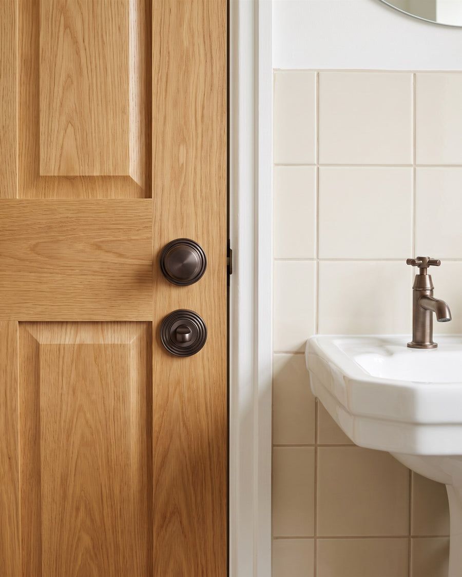 Matt bronze reeded knob above a matching reeded turn on an oak bathroom door, cream tiles and a bronze tap beyond