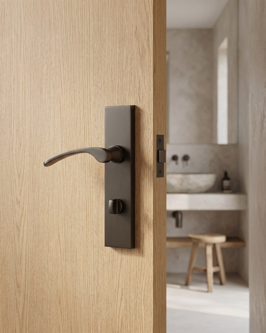 Matt bronze Sophia bathroom handle with lever and turn on a natural oak door, limewashed bathroom with stone basin beyond