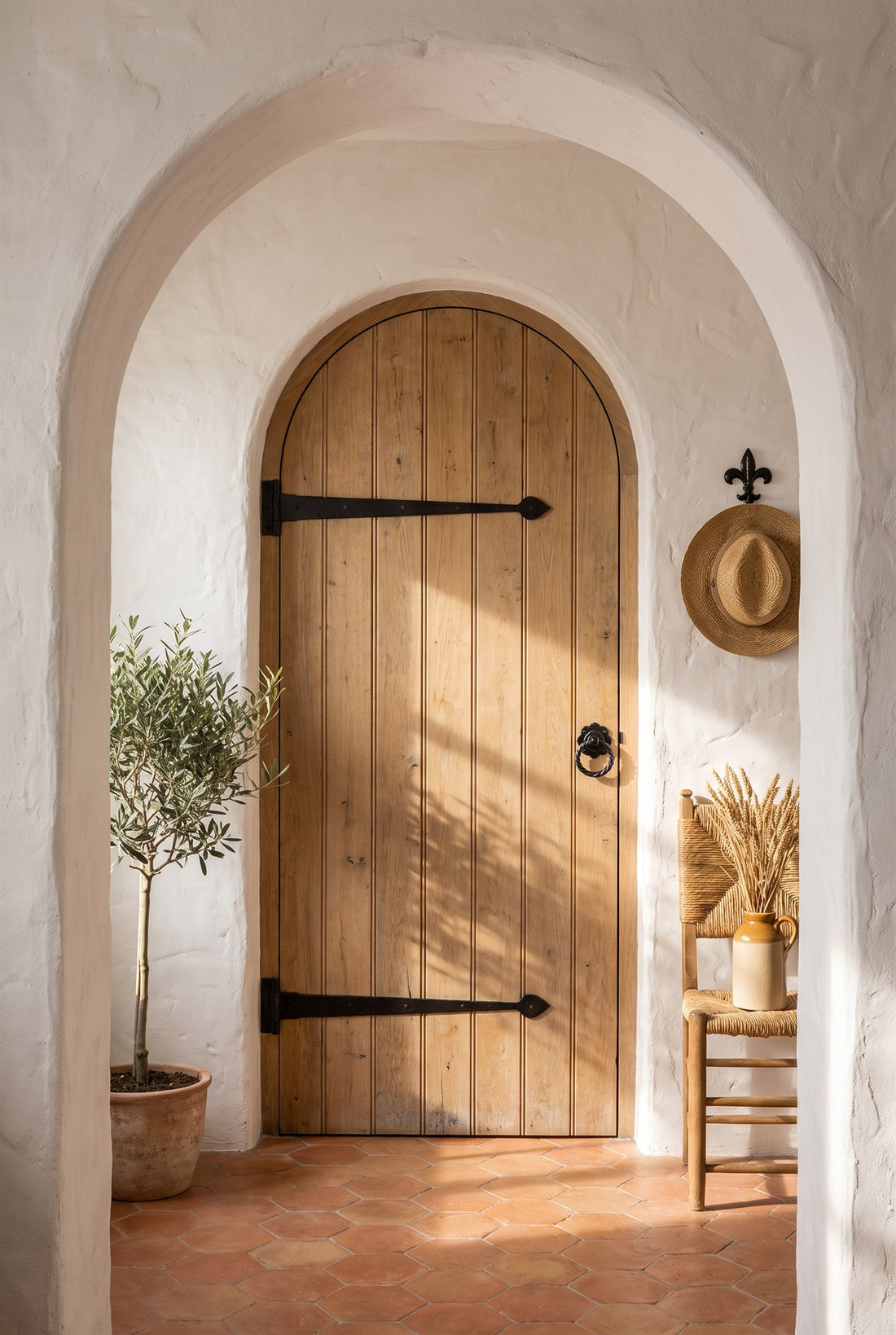 Mediterranean-style hall with white limewash, terracotta hexagon tiles and an olive tree, an arched oak plank door with black iron tee hinges and twisted ring handle, and a fleur-de-lys hook holding a straw hat