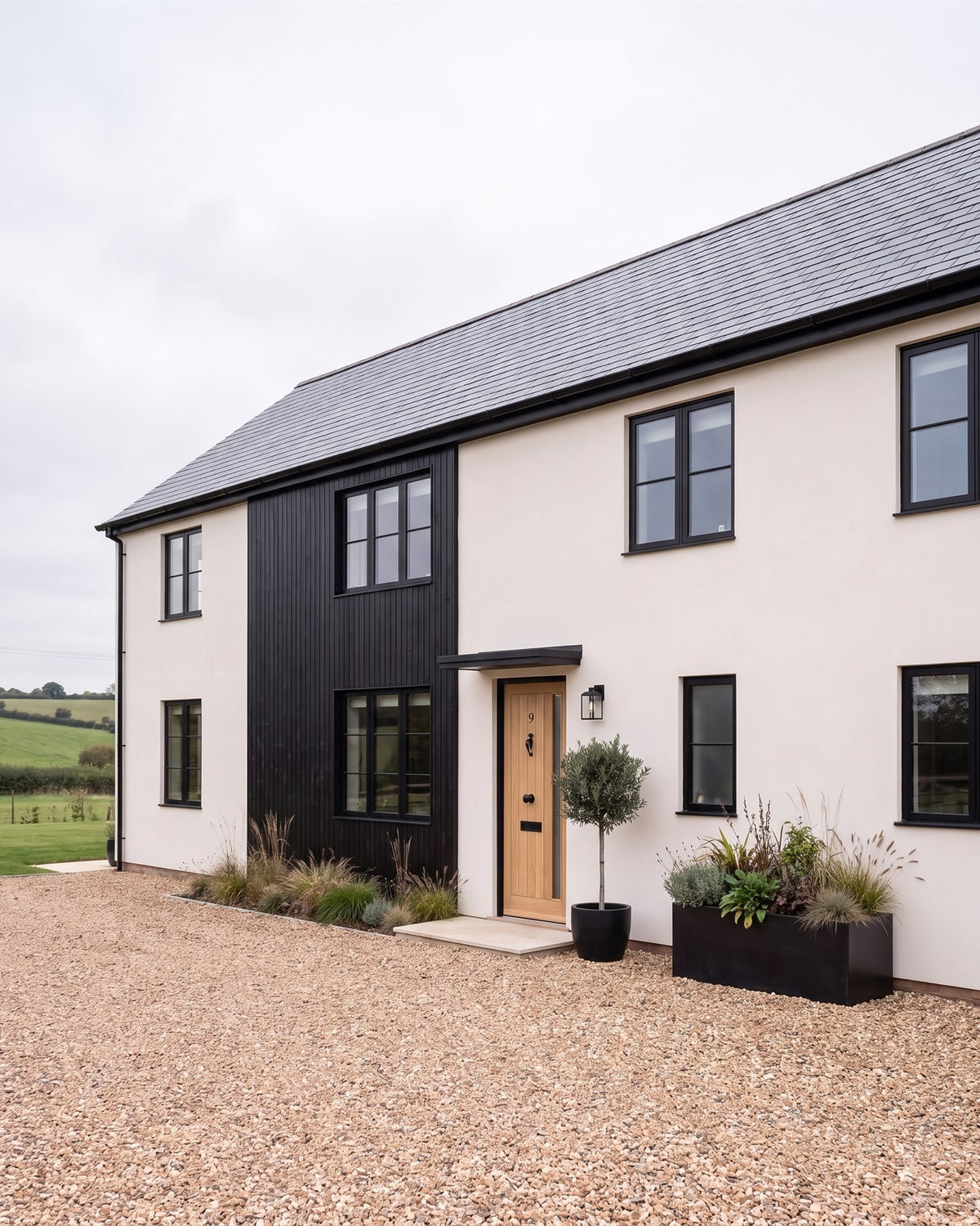 Modern farmhouse with white lime render, black timber cladding and black windows, an oak front door under a black canopy, gravel and ornamental grasses
