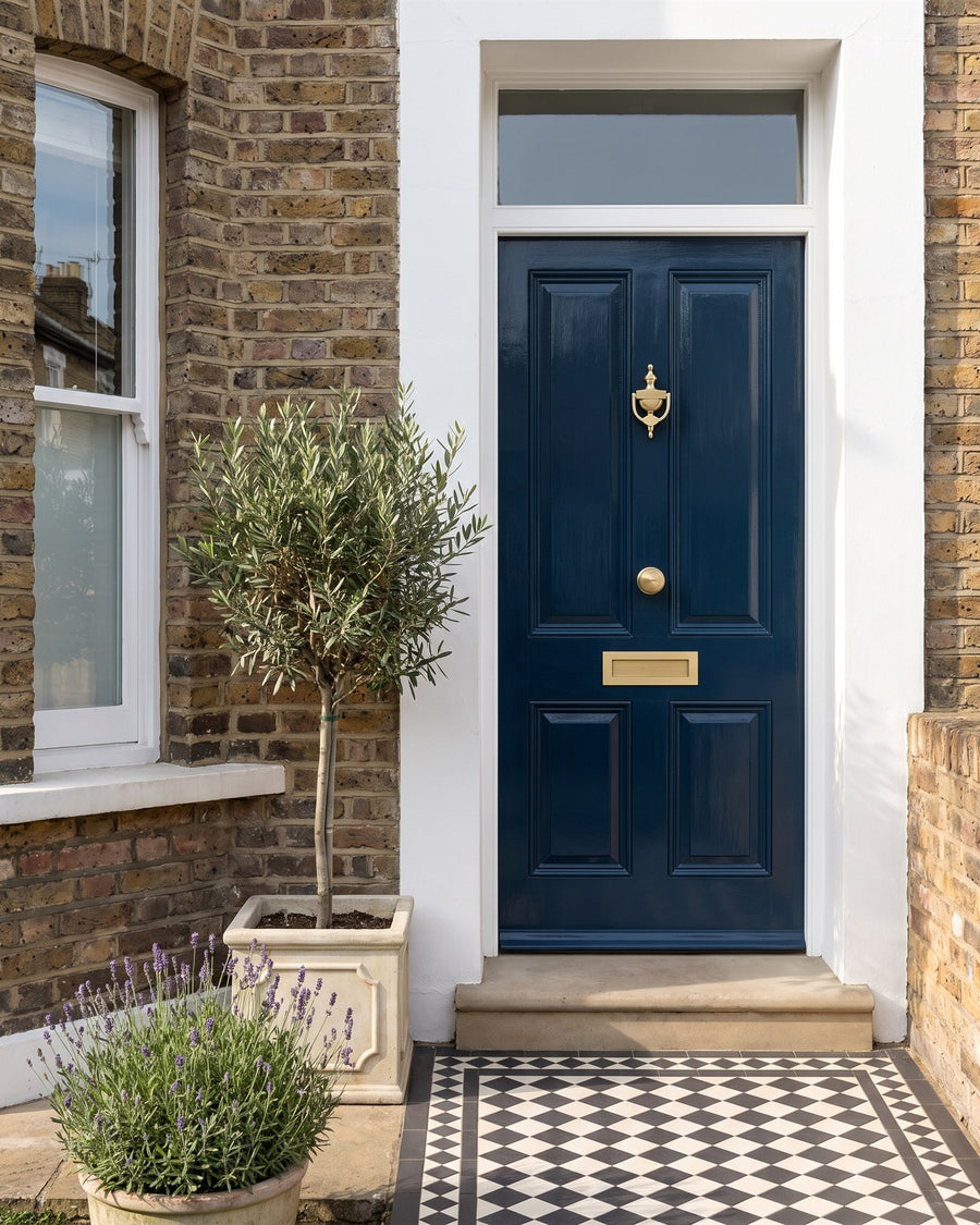 Victorian London terrace with a navy gloss front door, satin brass reeded knocker, knob and letter plate, white surround, black and white tiled path and a potted olive tree