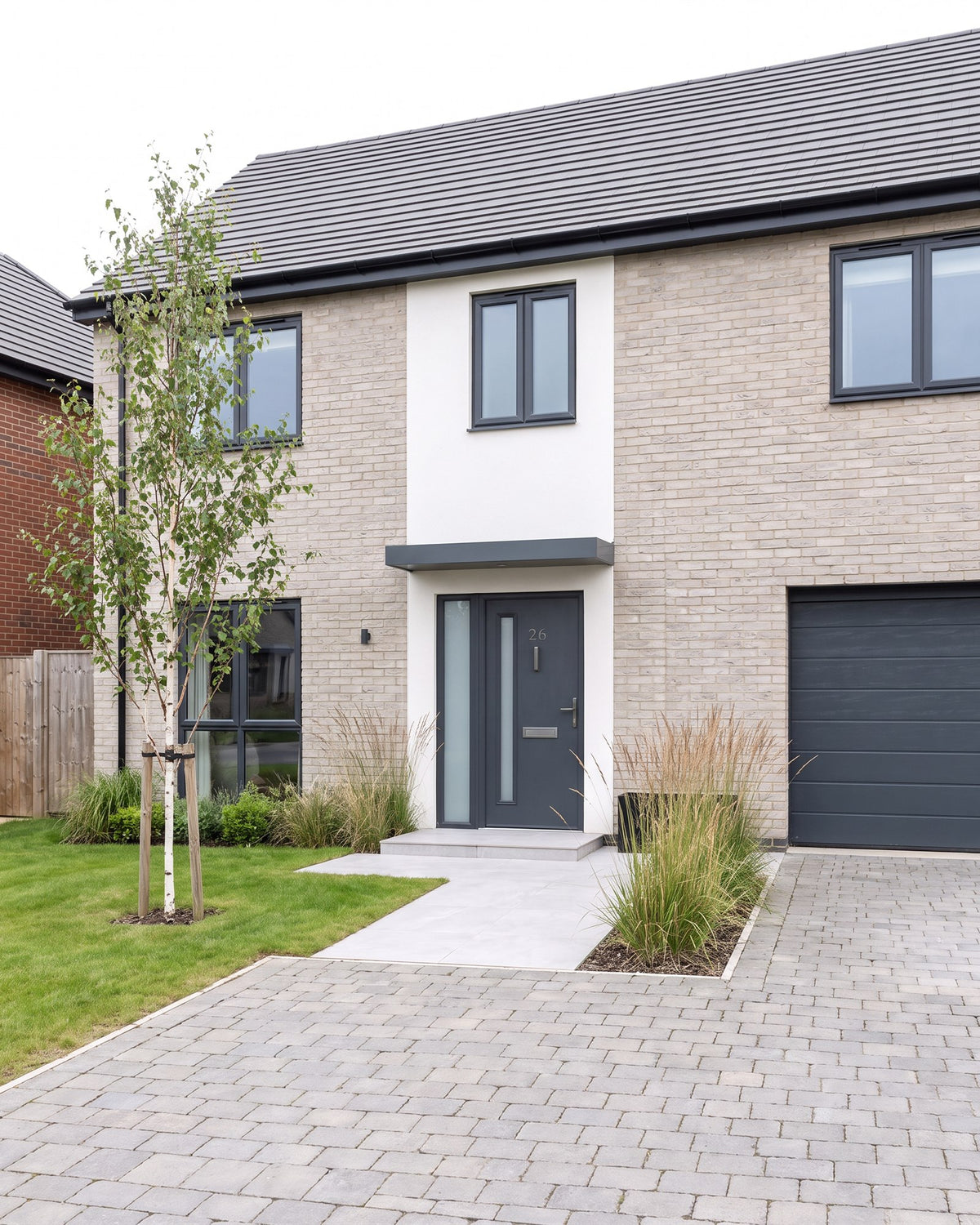 New-build detached house in pale buff brick with a white render entrance panel, anthracite windows and garage door, an anthracite composite front door under a flat canopy and ornamental grasses