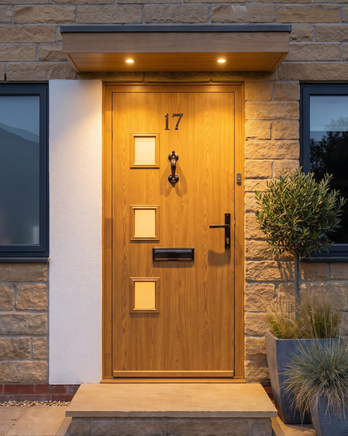 Golden oak composite door at dusk lit by canopy downlights, warm light through its three small windows