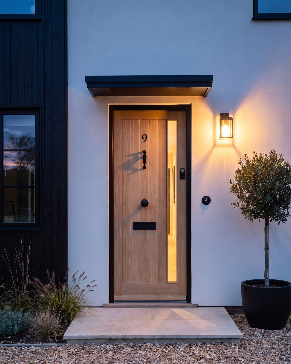 Oak front door with matt black fittings at dusk, lit by a black wall lantern and warm light through the glazed strip