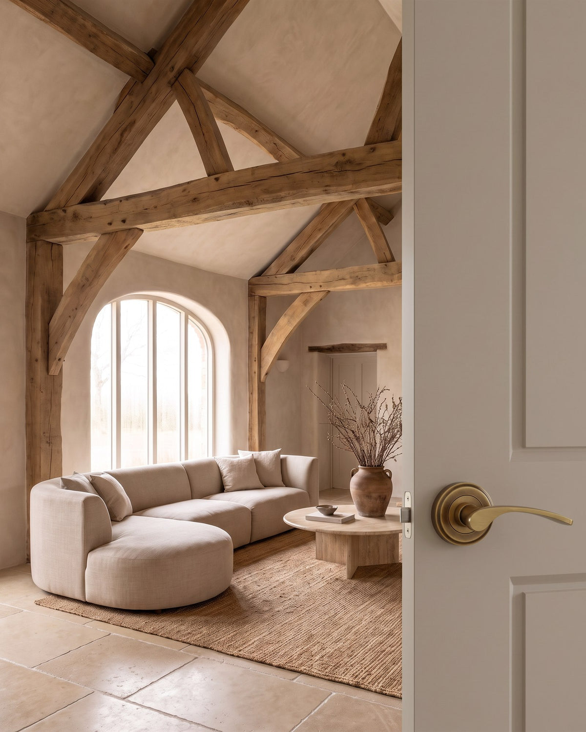 Organic modern barn living room with oak trusses and a curved sofa, seen past a mushroom door with an antique brass Sophia lever