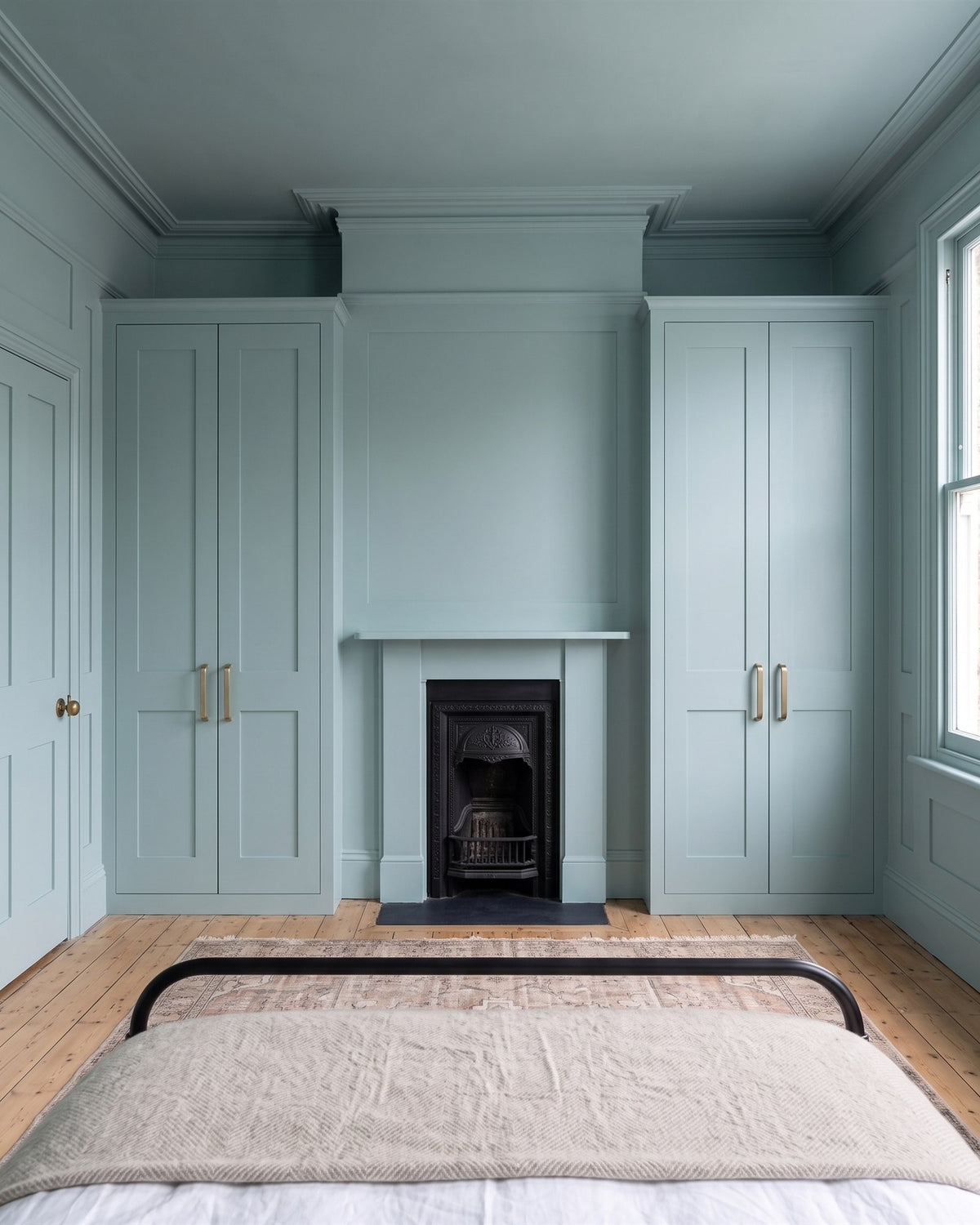 Colour-drenched Edwardian bedroom in soft blue-green panelling with a cast iron fireplace between two built-in wardrobes fitted with antique brass D handles, an iron bed in front