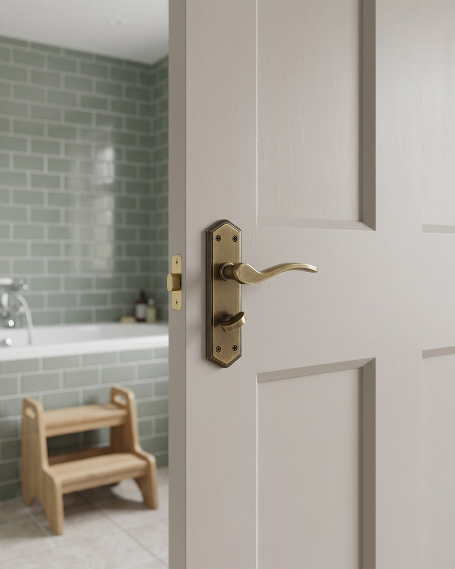 Paris antique brass and bronze bathroom handle with lever and turn on a soft greige panelled door, sage green tiled bathroom beyond