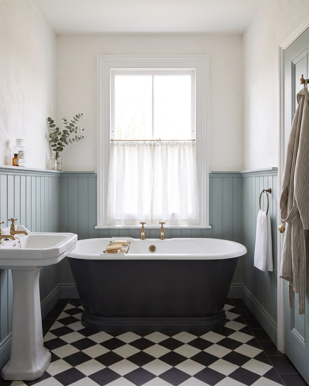 Period bathroom with a charcoal roll-top bath under a sash window with a cafe curtain, grey-blue tongue-and-groove panelling, a pedestal basin and a black and white floor