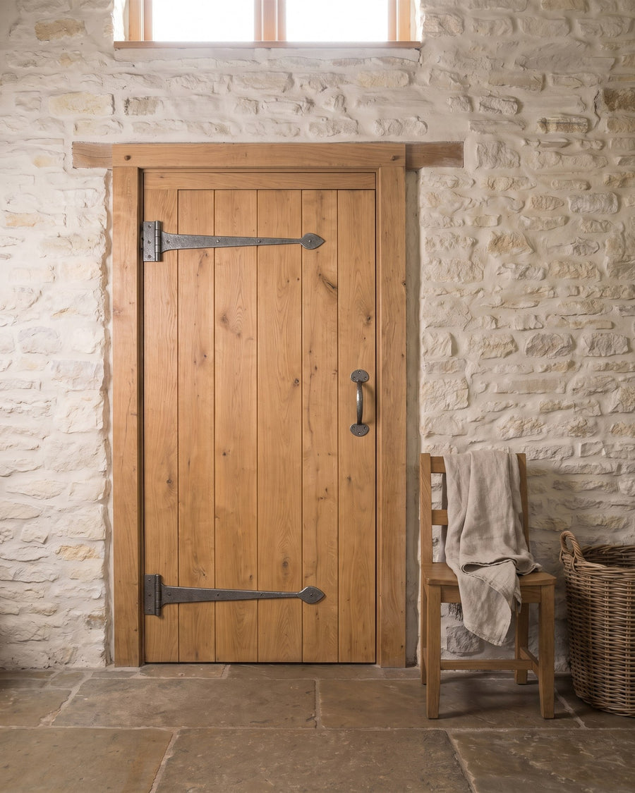 Oak ledged bedroom door in a limewashed stone wall with pewter tee hinges fixed to the frame and a pewter bow pull handle, beside an oak chair with a linen throw