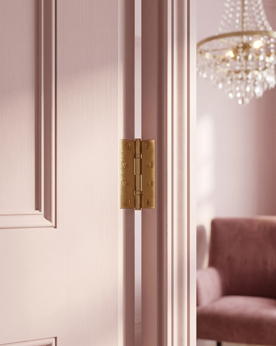 Polished brass butt hinge on the edge of a pink bedroom door with a chandelier and velvet chair blurred beyond