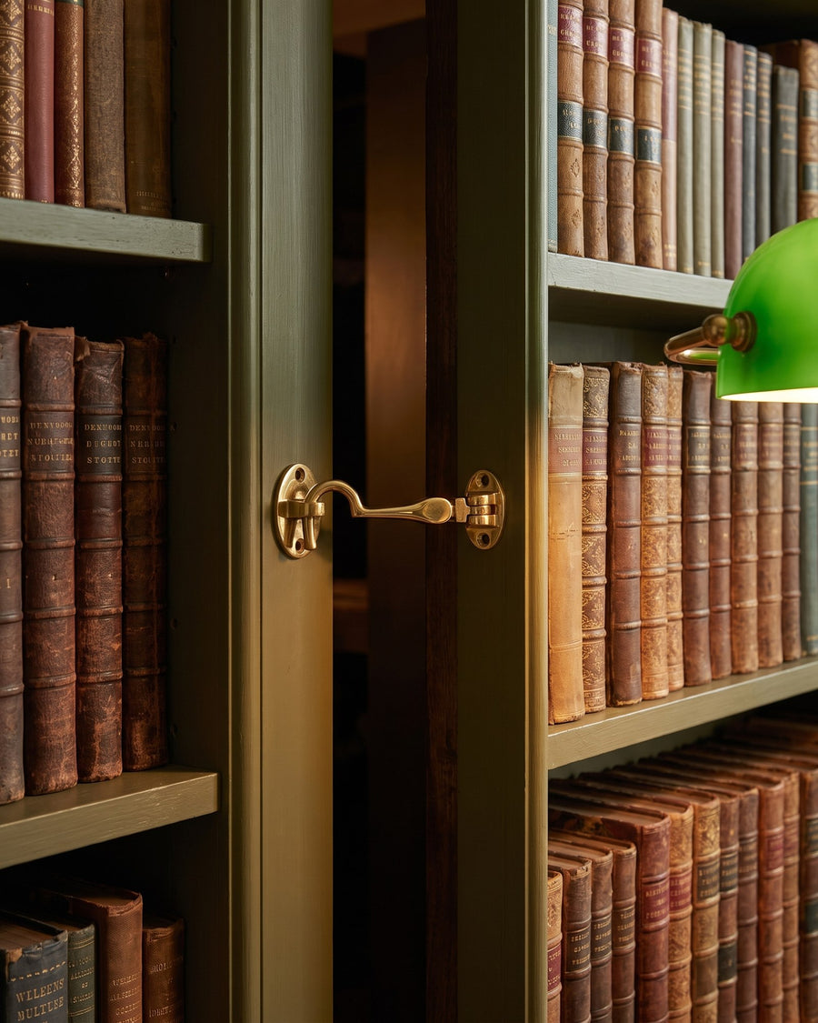 Polished brass cabin hook holding a hidden bookcase jib door slightly open between shelves of old books