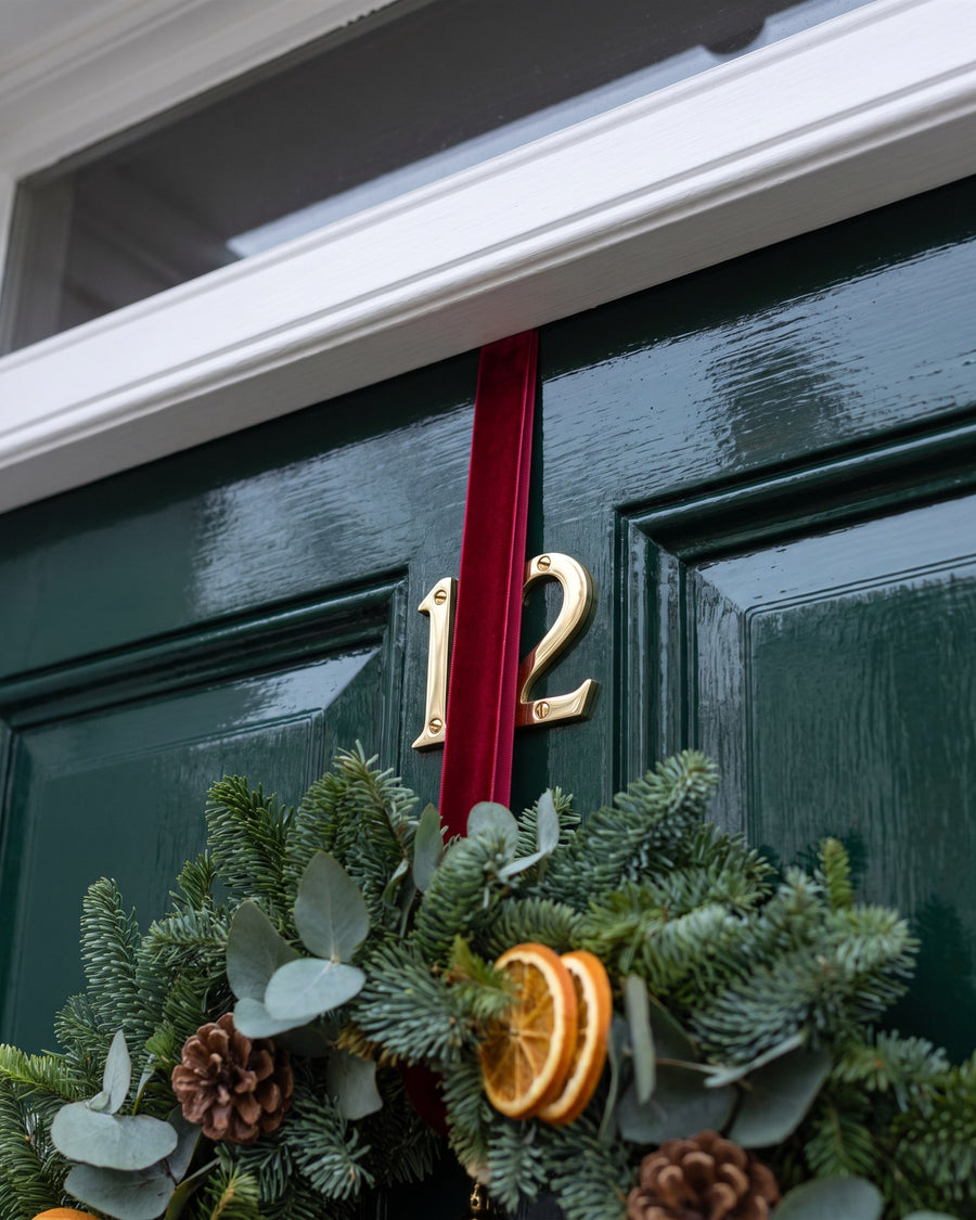 Polished brass screw-fixed numbers 1 and 2 on a bottle-green door with the red velvet wreath ribbon running down past them