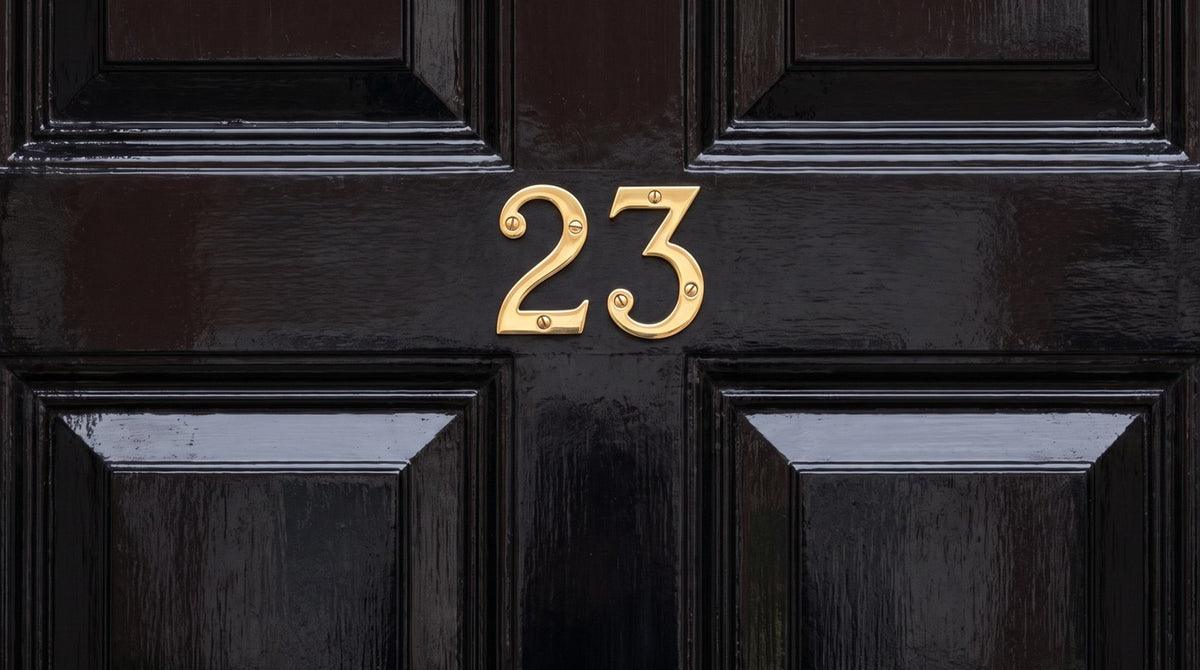 Polished brass serif house numbers 23 screw-fixed to the top rail of a glossy black panelled front door