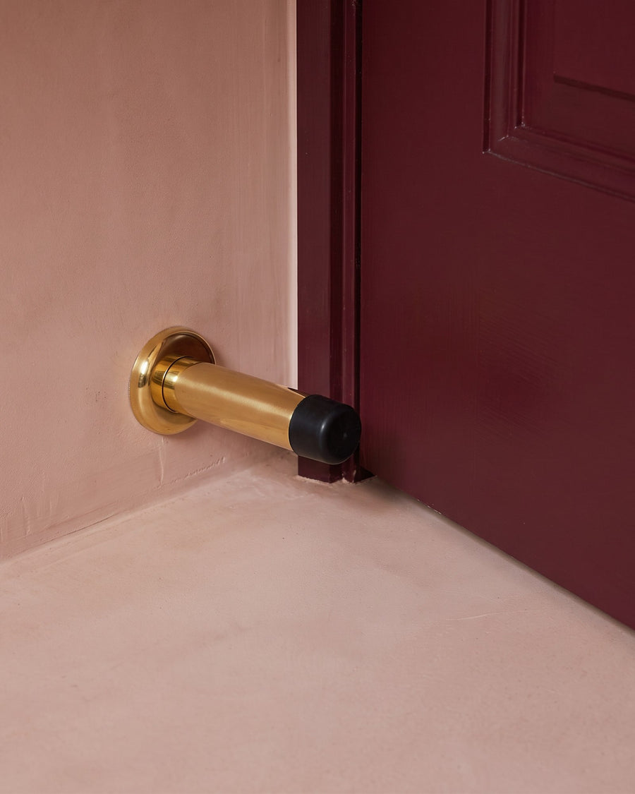 Burgundy door resting against a polished brass projection door stop on a blush micro-cement wall