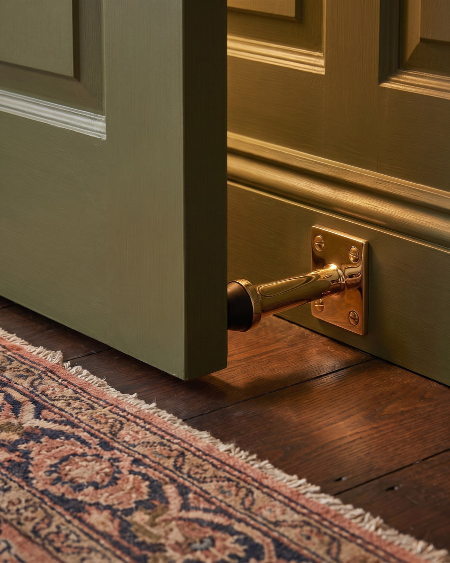 Olive panelled door resting against a polished brass projection door stop on the skirting above a Persian rug