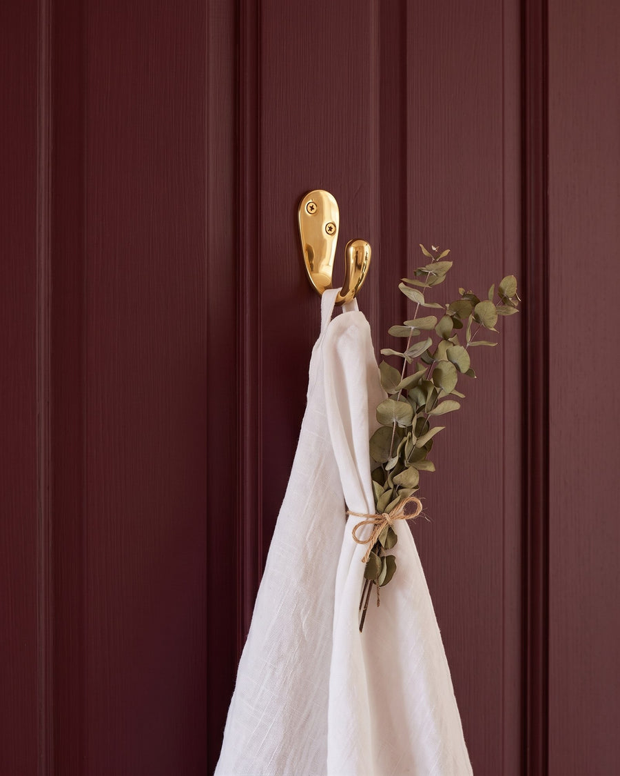 Polished brass robe hook holding a white linen towel and a bundle of dried eucalyptus on a burgundy door