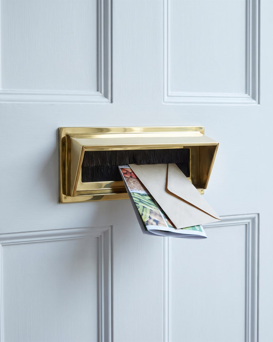 Polished brass letter tidy with a brush draught excluder on the inside of a white door, an envelope and seed catalogue dropping through