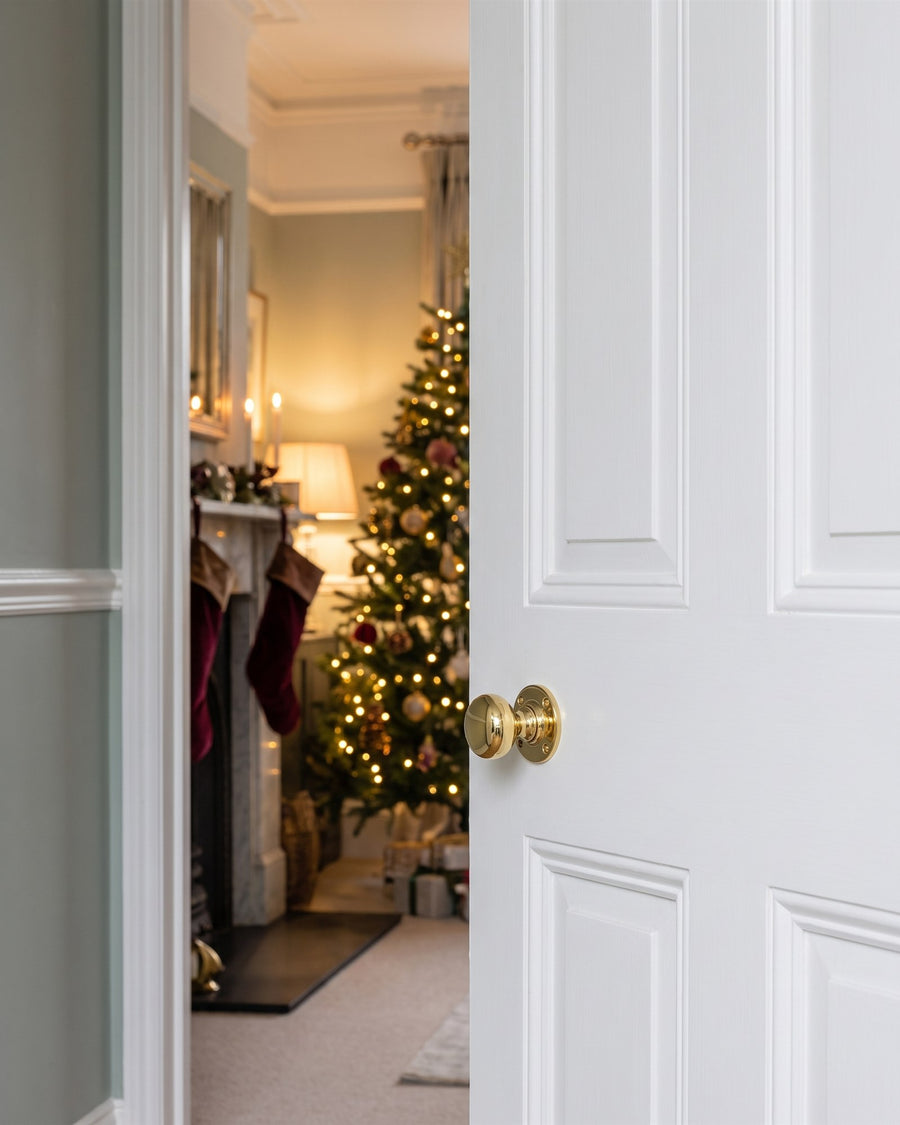 Plain round polished brass Victoria knob on a white four-panel drawing room door, open onto a fireplace hung with velvet stockings and a lit Christmas tree