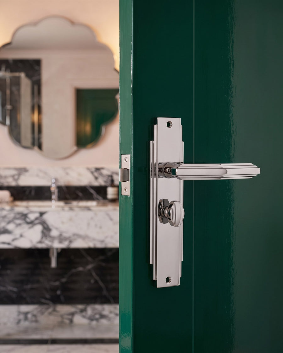 Polished chrome Art Deco bathroom handle on a stepped plate with turn, on a high-gloss emerald door, marble bathroom beyond