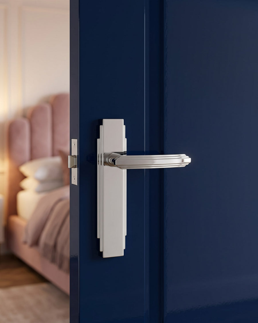 Polished chrome Art Deco latch handle on a stepped plate on an ink navy lacquered bedroom door, pink velvet bed beyond