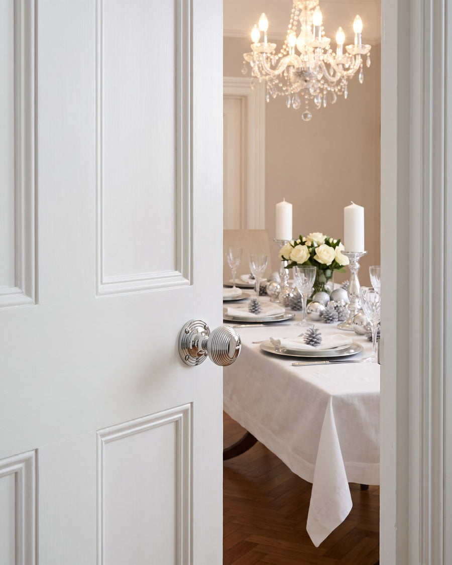 Polished chrome reeded beehive knob on a white panelled door opening onto a dining table laid with white linen, silver chargers and candles under a crystal chandelier