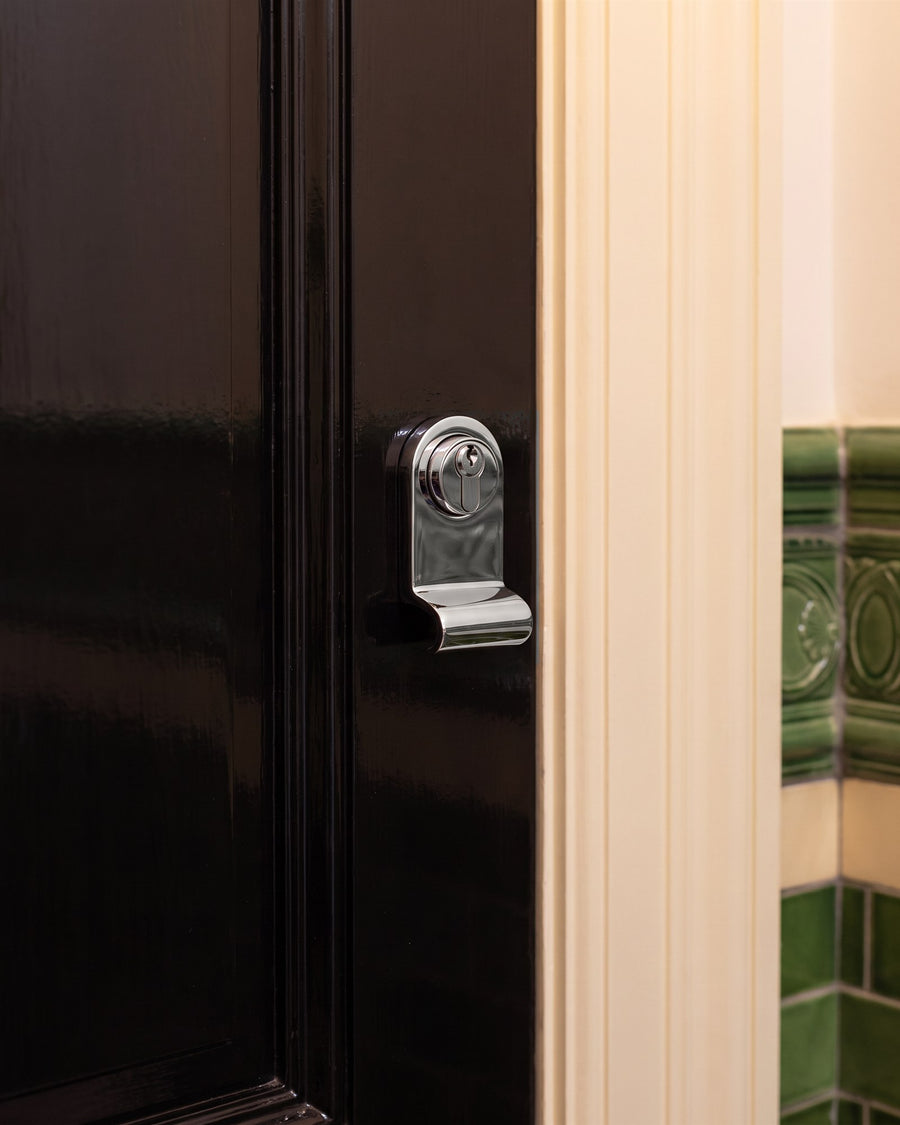 Polished chrome cylinder pull around a euro cylinder on a black flat door beside a cream architrave and green glazed corridor tiles