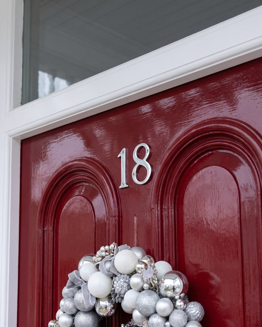 Polished chrome numbers 18 between two round-arched panels on a red front door, the top of a silver and white wreath below