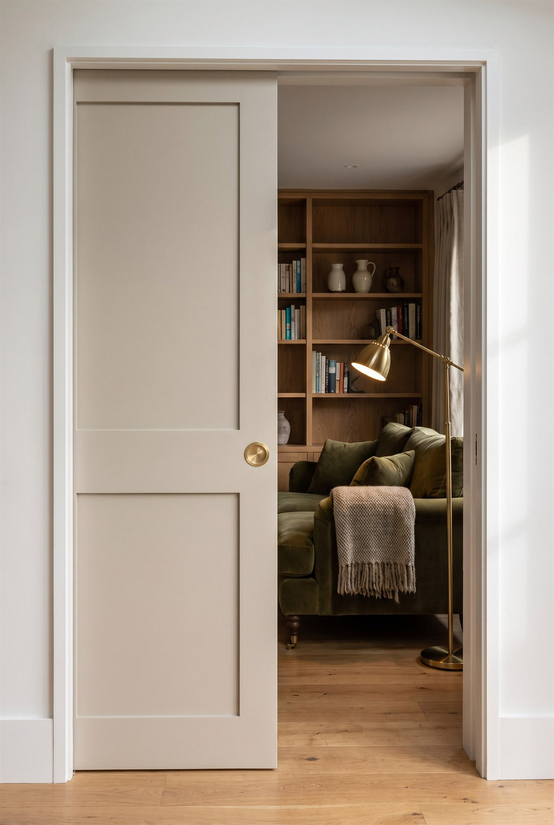 Putty painted two-panel pocket door half slid into the wall with a circular satin brass flush pull, opening onto a snug with an olive velvet sofa and oak shelves
