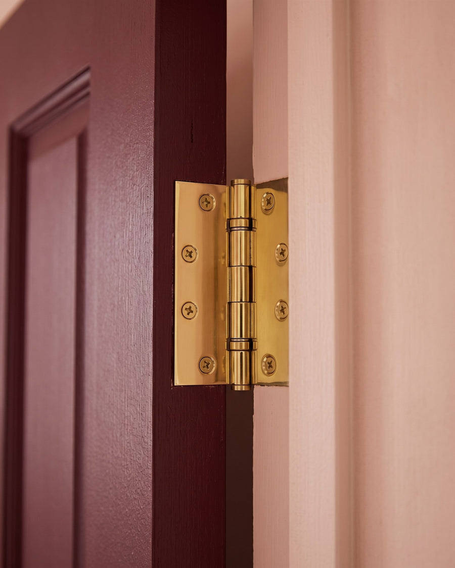 Bright PVD brass ball-bearing butt hinge joining a deep burgundy door to its frame