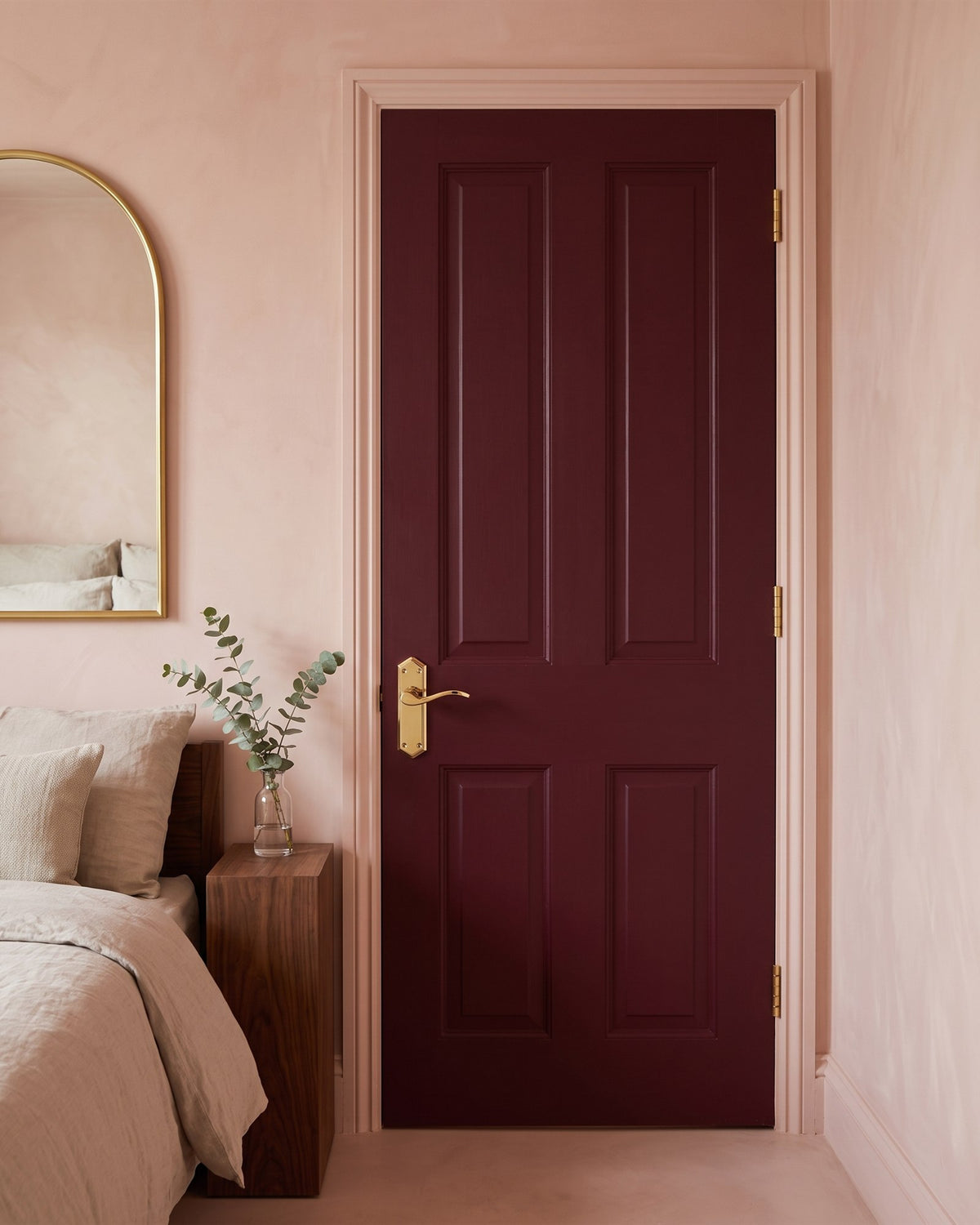 Deep burgundy bedroom door with a PVD brass Paris scroll lever latch handle, beside a linen bed and walnut bedside table