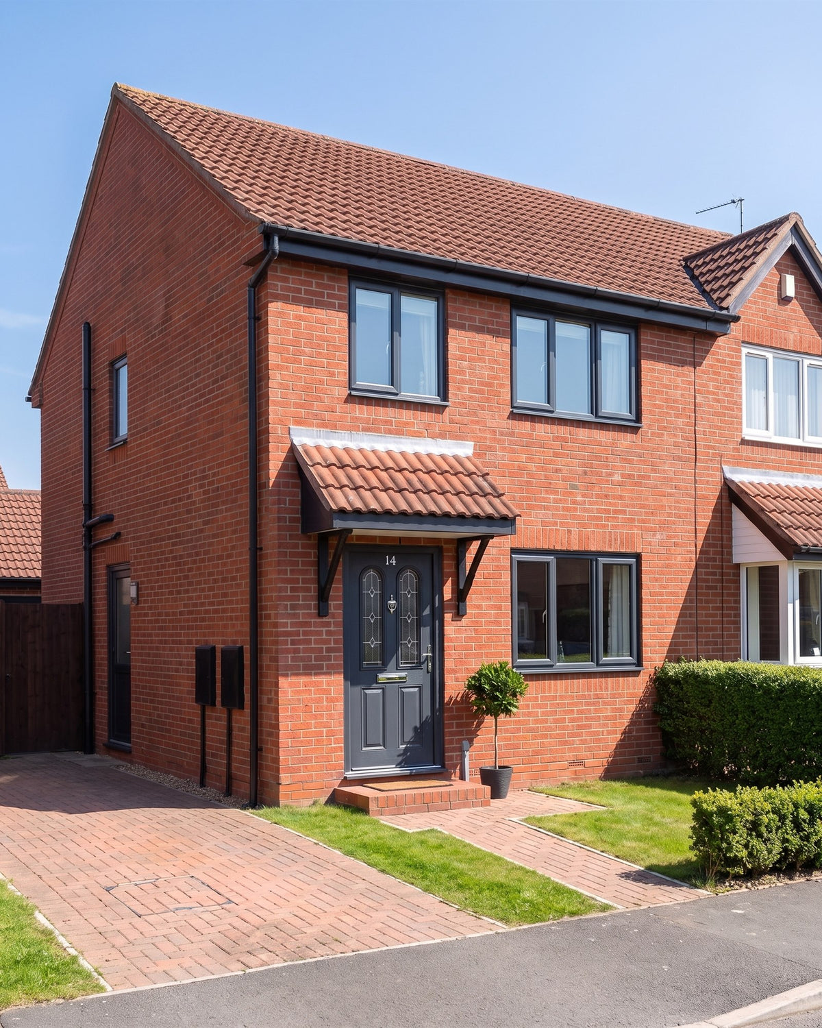 1990s red-brick semi-detached house with anthracite windows, a tiled canopy porch over an anthracite composite front door, a lawn and block-paved path
