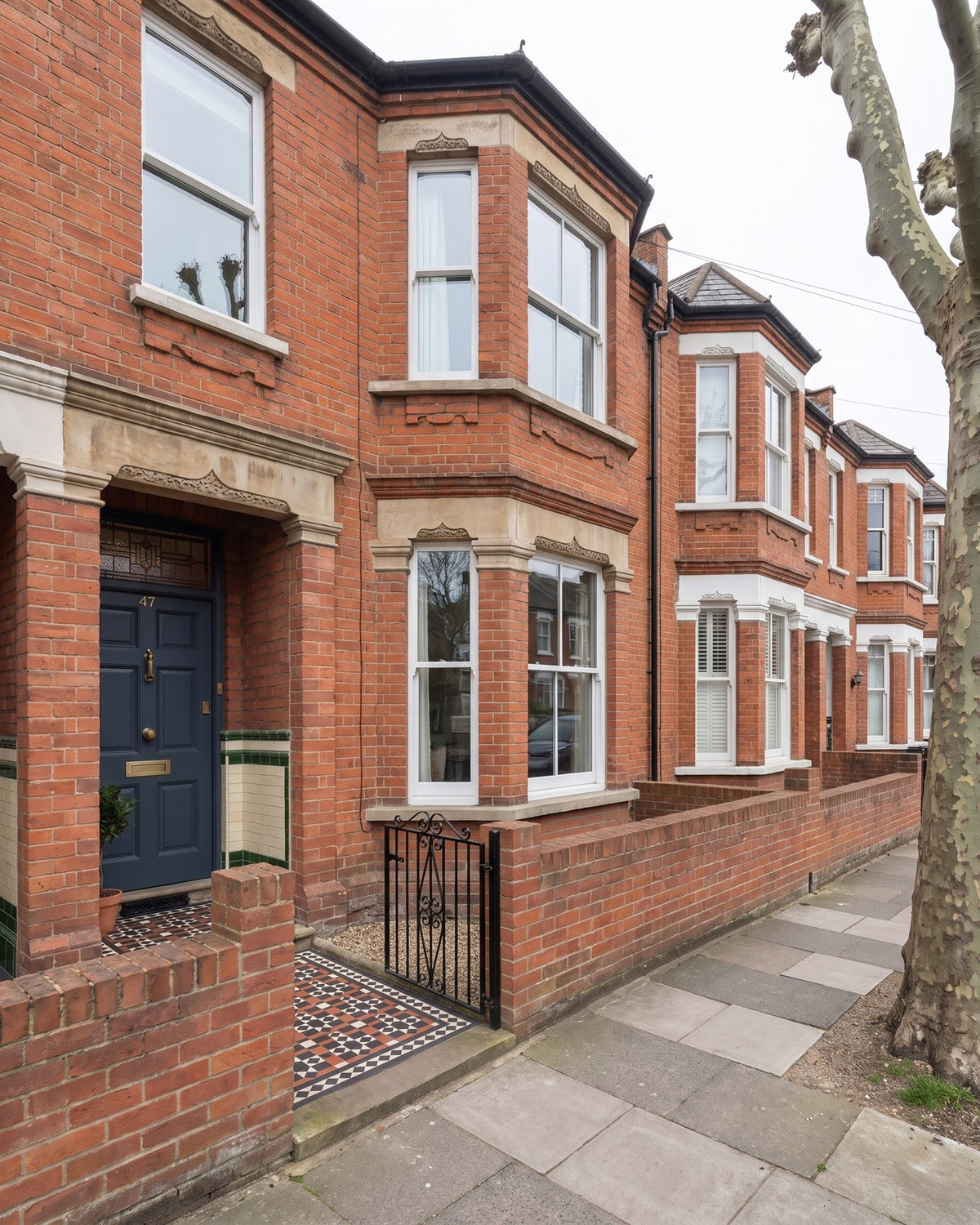 Red-brick Victorian terrace with a two-storey canted bay, low front wall and iron gate, an encaustic tiled path leading to a grey front door in a recessed tiled porch