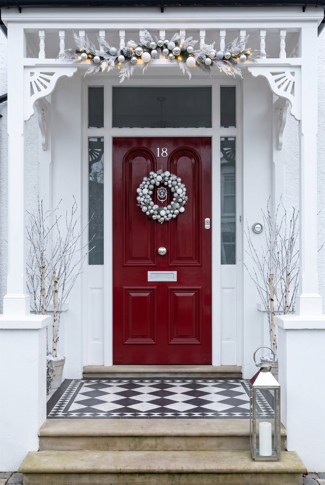 Glossy red Edwardian front door in a white fretwork porch dressed with a silver and white wreath and frosted swag, fitted with polished chrome numbers 18, lion knocker, stepped centre knob, letter plate, cylinder pull and china bell push