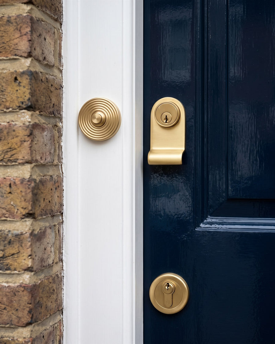 Satin brass reeded bell push on a white door surround beside a satin brass cylinder pull and round euro escutcheon on a navy front door