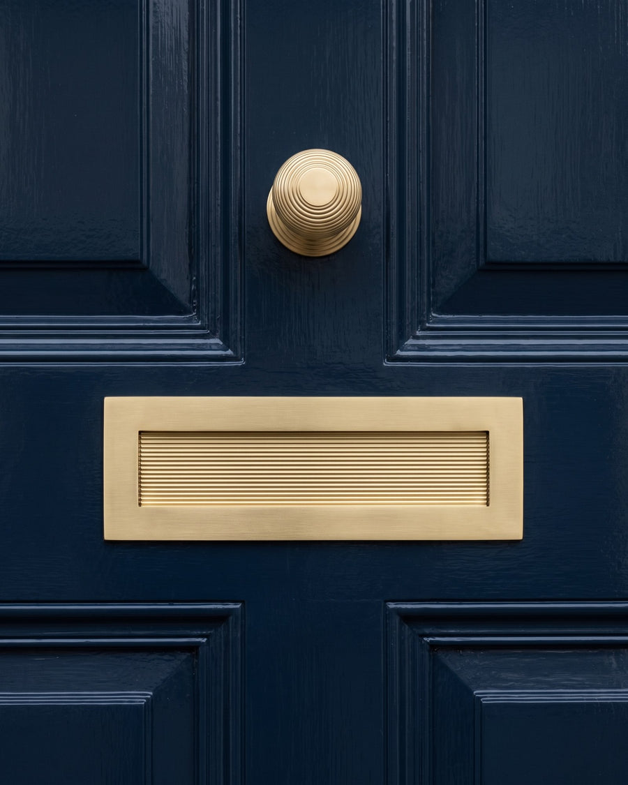 Satin brass reeded letter plate on the middle rail of a navy four-panel door, with the reeded centre knob above