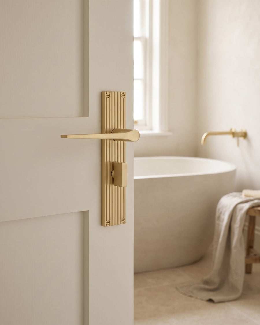 Satin brass Gio reeded bathroom lever with thumbturn on a warm white door, freestanding stone bath beyond
