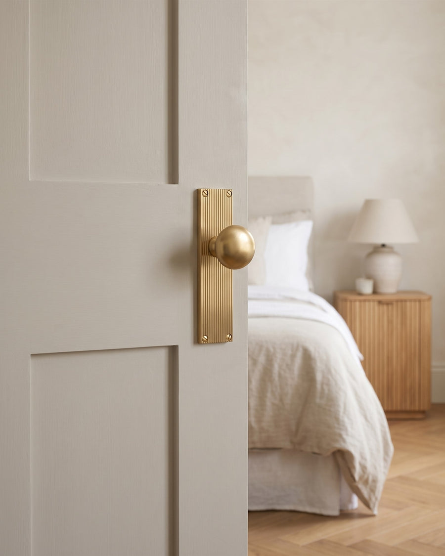 Satin brass Victoria knob on a reeded backplate on a stone painted bedroom door, linen bed and fluted oak bedside table beyond