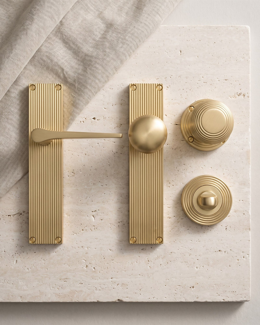 Flat-lay of satin brass reeded fittings on travertine: lever on reeded plate, knob on reeded plate, reeded mortice knob and reeded turn and release