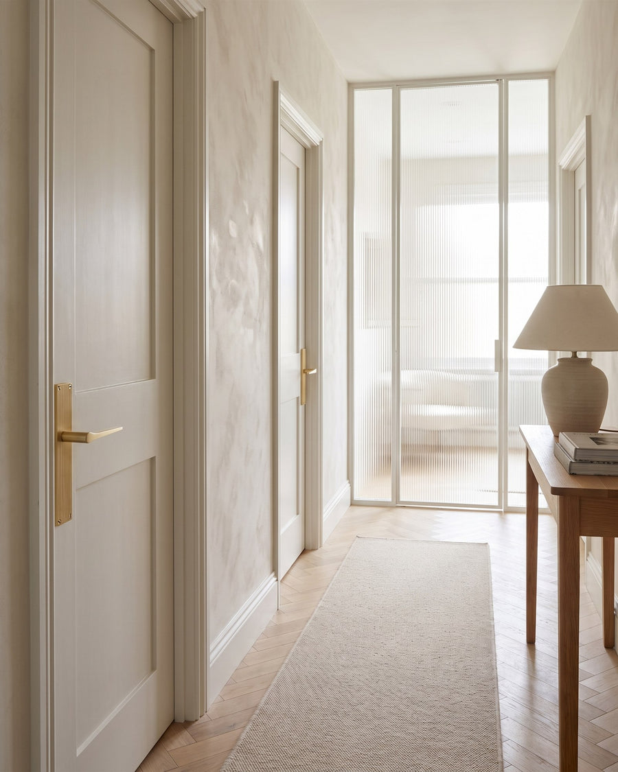 Limewashed landing with warm white Shaker doors, satin brass reeded lever handles, oak herringbone floor and a fluted glass screen