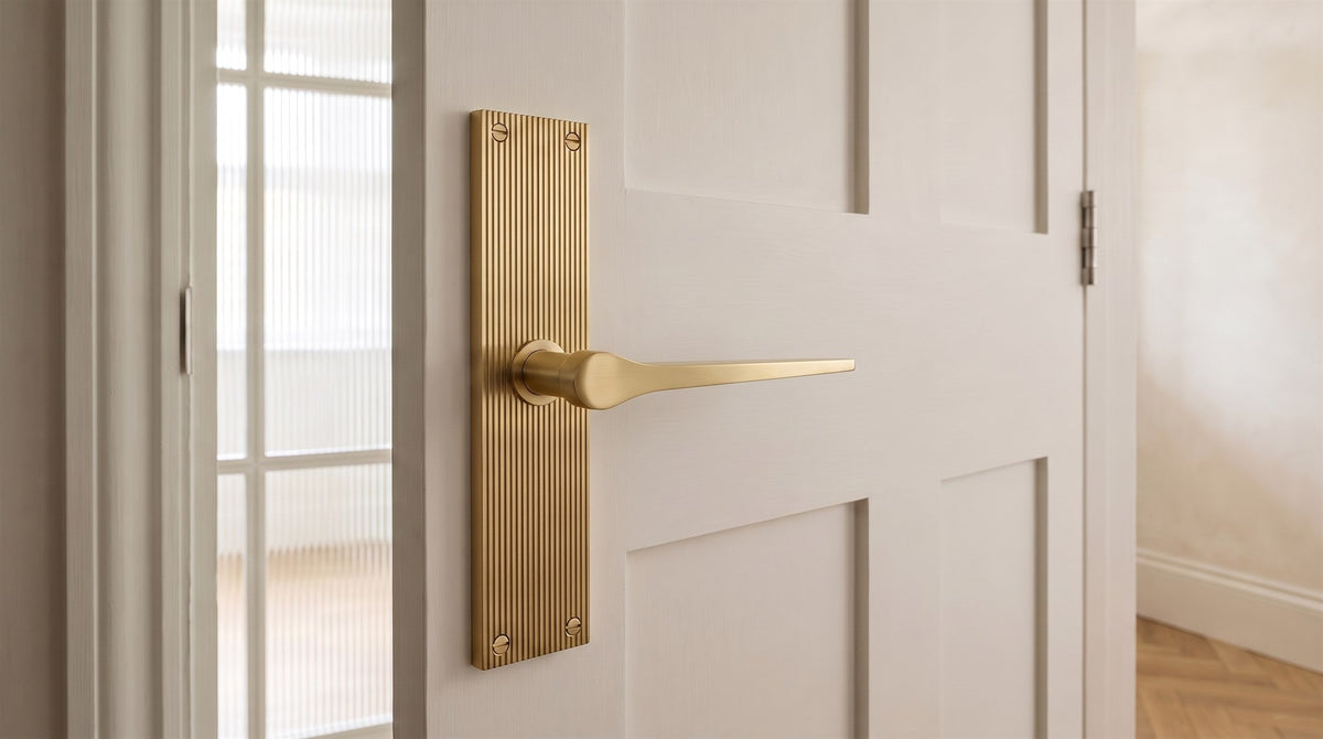 Satin brass Gio reeded lever handle on a warm white Shaker door, with a fluted glass screen and oak herringbone floor behind