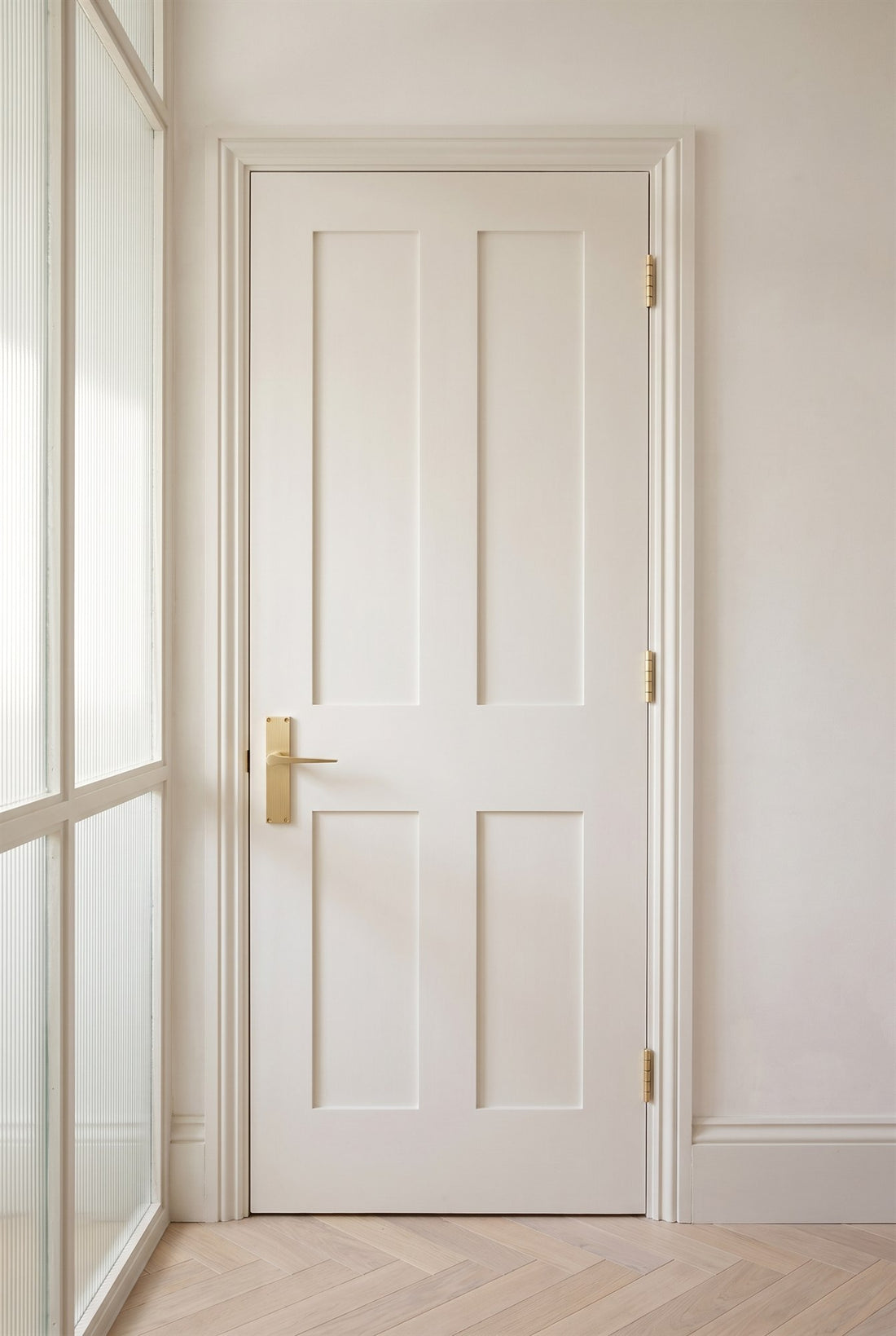 Warm white four-panel Shaker door with a satin brass Gio reeded lever handle and satin brass butt hinges, fluted glass screen alongside