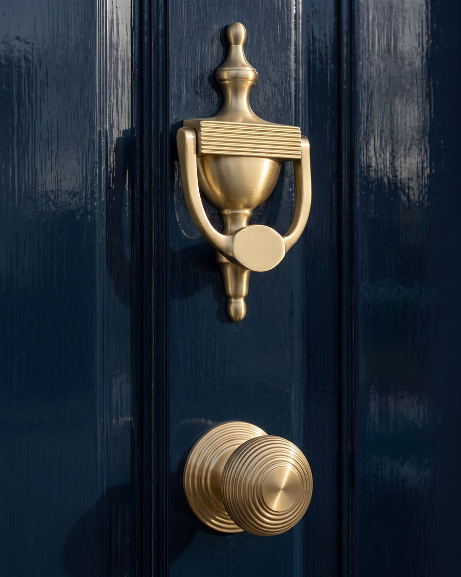 Satin brass reeded urn knocker above a reeded centre door knob on a navy gloss door, low light showing the ribbed detail