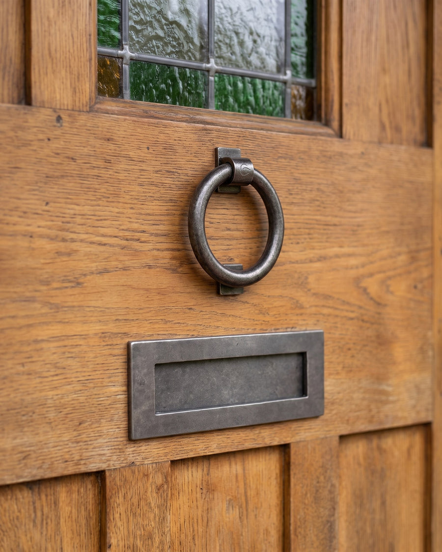 Rustic pewter ring knocker above a rustic pewter letter plate on a weathered oak door below a green leaded light