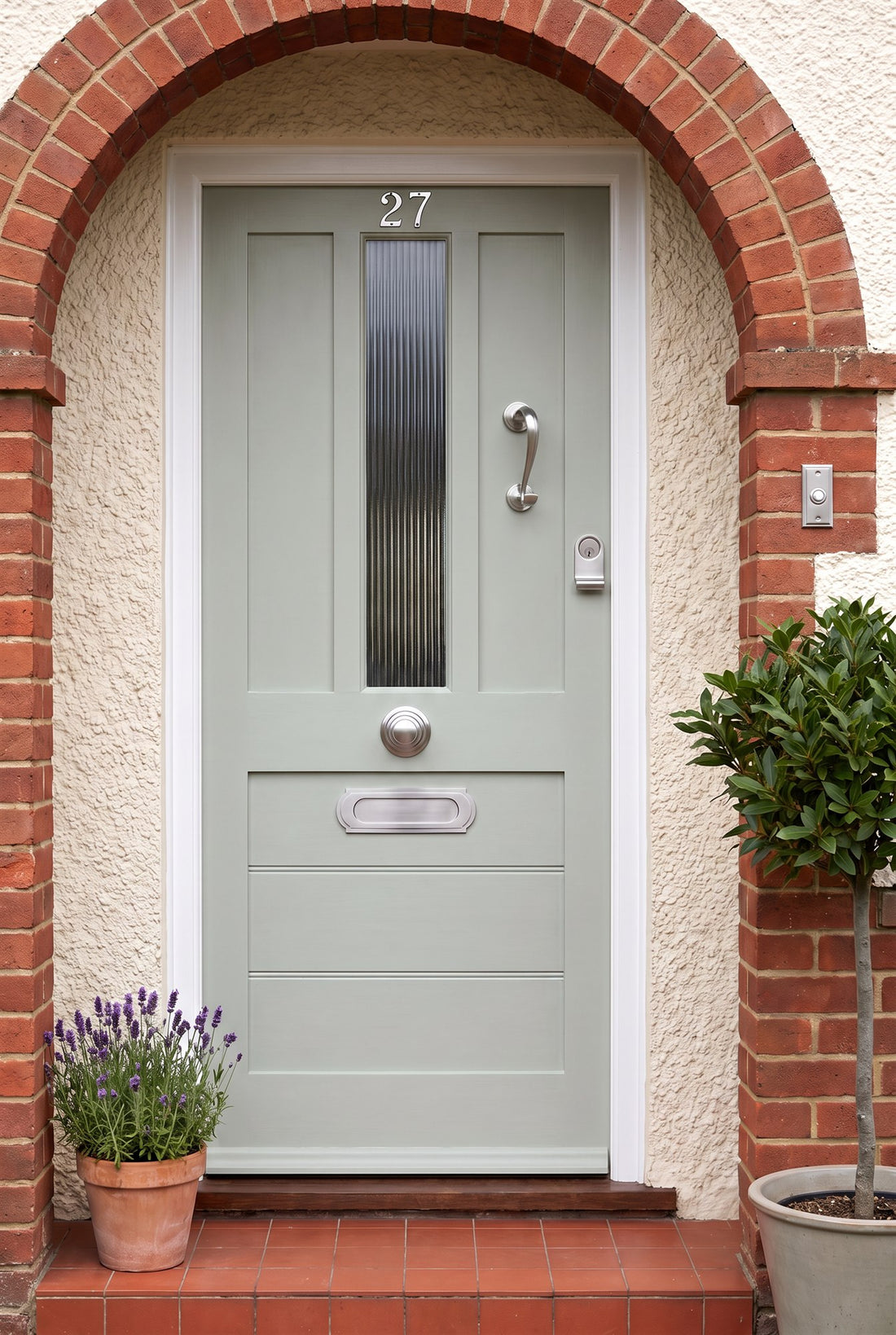 Soft sage green 1930s front door with a reeded glass panel under a brick porch arch, fitted with satin chrome number 27, Sloane scroll knocker, Sloane centre knob, Sloane letter plate, cylinder pull and stepped bell push
